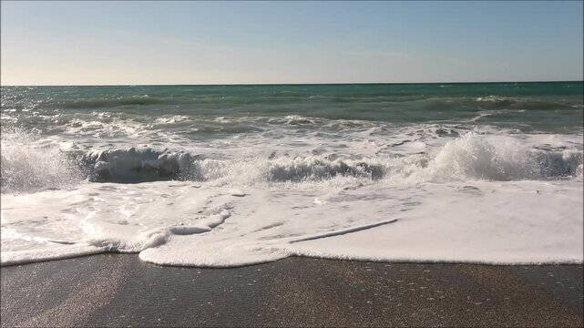 Powerful ocean waves crash onto a sandy beach under a clear blue sky