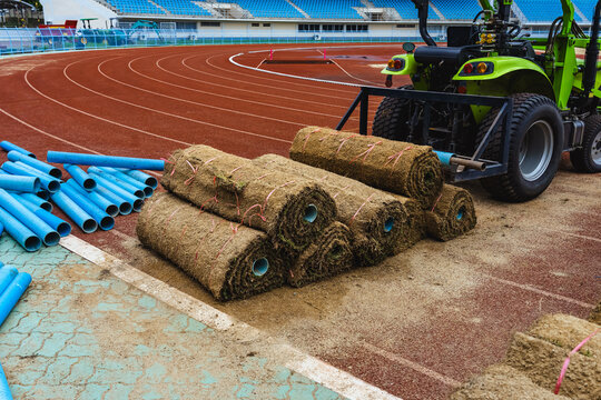 Landscaping tractor preparing ground for new grass rolls at sports complex. Professional sports field renovation featuring natural sod rolls. Installation of fresh natural turf rolls at large stadium.
