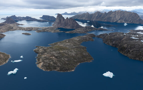 Aerial view of rugged mountains and rocky islands surrounded by deep blue fjords with floating icebergs under an overcast sky Greenland.
