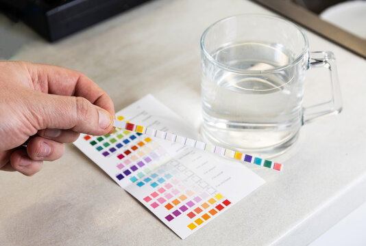 Hand holding water quality test strip displaying colored chemical indicators for comparison with reference chart, transparent glass mug of water next to paper results table