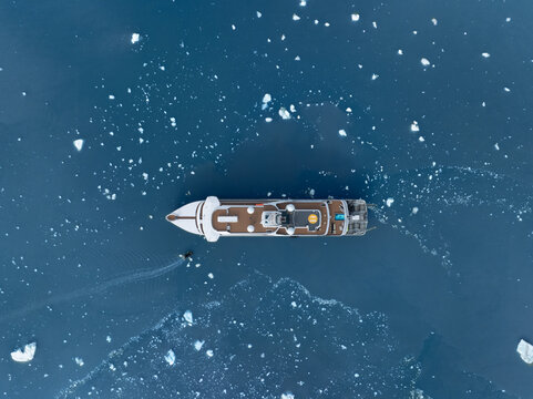Aerial view of an expedition cruise ship and a small boat surrounded by floating ice fragments in the blue sea in Greenland.