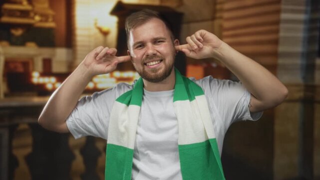 Man wearing green and white scarf plugs ears with fingers and smiles inside a church building, caucasian young fan in white tshirt; defiance.