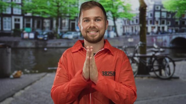 Man in orange prisoner jumpsuit numbered 38258 with palms pressed together praying beside a canal on an amsterdam street near parked bicycles and a bridge; hopeful plea redemption.