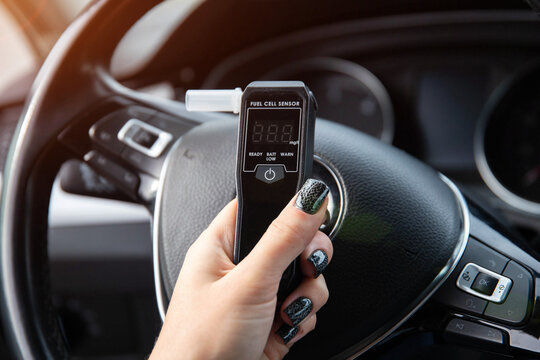 Woman's Hand Holding Breathalyzer Over Steering Wheel. Female Driver Checks Sobriety Before Trip.