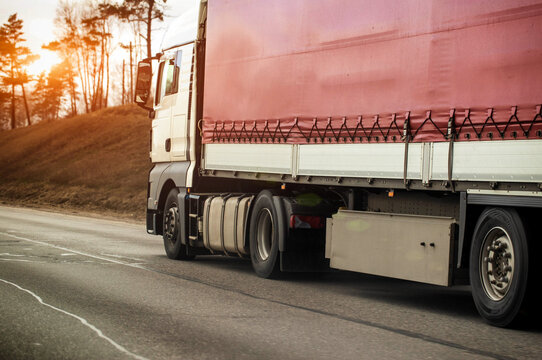 Heavy white cargo truck with red tarp trailer transports goods along asphalt highway, wheels show motion blur, scene features bare trees and cloudy sky