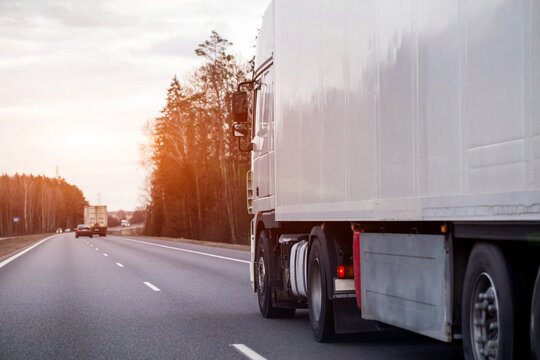 White semi-trailer truck travels highway, side view trailer, wheels, forest, overcast sky, depicting cargo transport logistics industry efficiency, evening sunset. Copy space for text