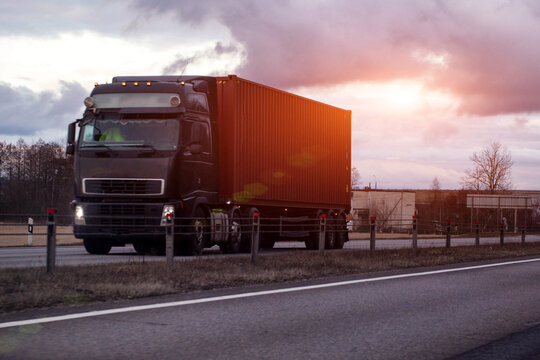 Semi-truck carrying red shipping container on highway under cloudy sky at dusk or dawn, illustrating cargo transportation and logistics industry. Copy space for text