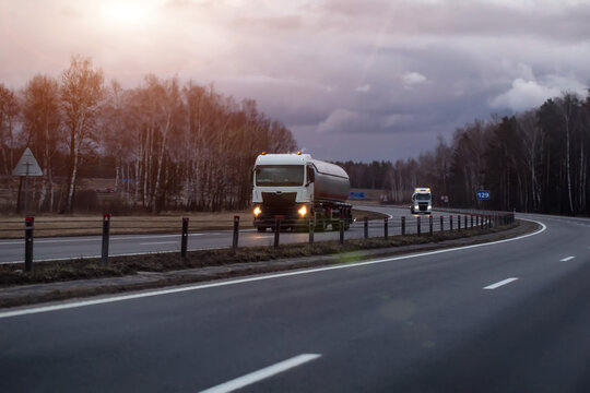 Tanker truck with illuminated headlights travels on curved highway through barren landscape under cloudy evening sky, another truck in distance, evening sunset. Copy space for text