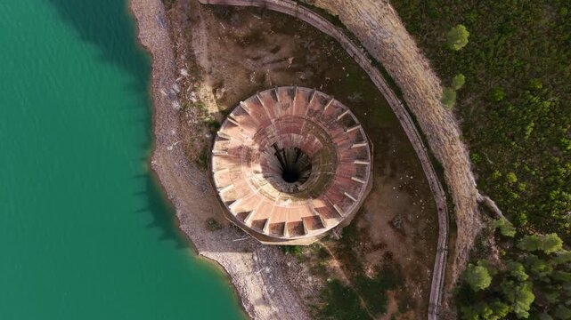 Top-down aerial of reservoir intake tower with bell-mouth spillway, radial buttresses and shaft drop inlet, showing hydraulic intake structure at low water level shoreline