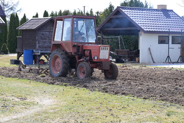 Rolnik w czerwonym ciągniku orze, sprężynuje glebę, ziemię w ogródku przydomowym na wsi © Joanna