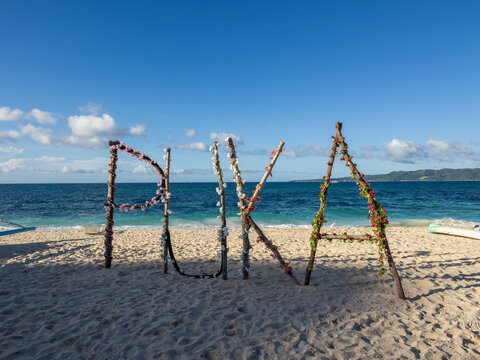 The iconic 'PUKA' sign, adorned with vibrant blossoms on driftwood, stands on the pristine sands of Puka Beach in Boracay, Philippines. The shimmering turquoise ocean stretches to the horizon.