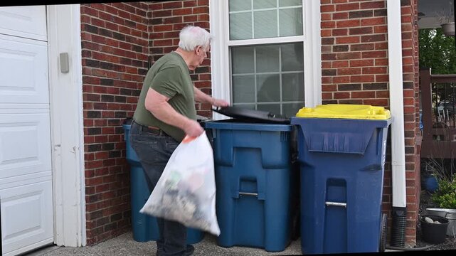Older white man approaches blue trash bins and places white plastic garbage bag in the center bin and closes the lid.
