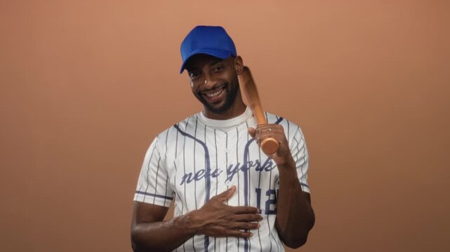 Man in a striped baseball jersey holding a wooden bat and smiling in a peach studio; joyful confidence.