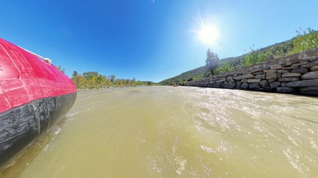 A low-angle shot of a rafting boat cutting through fast-moving water, with the sun high in the clear blue sky, surrounded by lush trees and a stone wall on one side of the river.