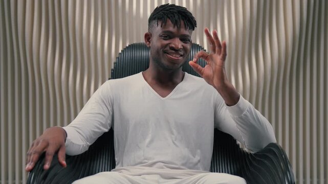 Futurism African American man showing ko gesture sitting in chair in modern futuristic space room studio smiling joyful glad satisfied male guy happy with modern technology fine reaction good quality