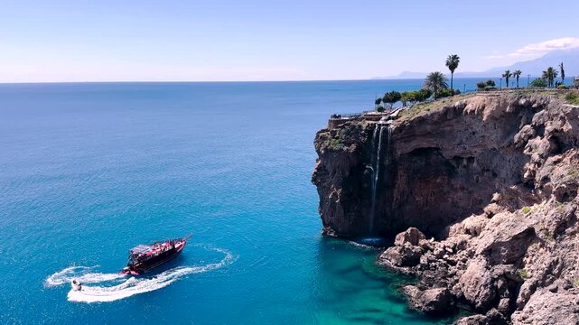 Scenic Antalya T&uuml;rkiye coast with dramatic cliffs and a small waterfall flowing into the Mediterranean. A tour boat and speedboat move across turquoise water under a clear blue sky.