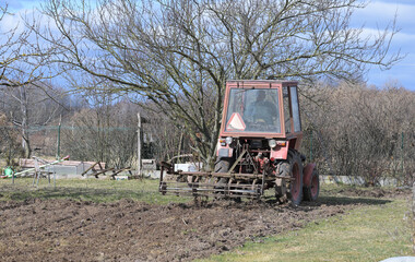 Rolnik w czerwonym ciągniku orze, sprężynuje glebę, ziemię w ogródku przydomowym na wsi © Joanna