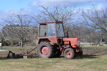 Rolnik w czerwonym ciągniku orze, sprężynuje glebę, ziemię w ogródku przydomowym na wsi © Joanna
