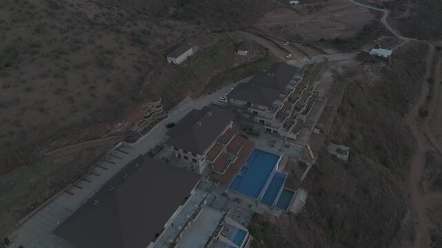 Aerial view of the Aravalli hills near Udaipur, Rajasthan at sunset, showing winding roads, patchwork agricultural fields, native trees, and scattered village houses across rugged terrain.