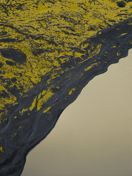 Aerial view of a red off-road vehicle driving on a dark gravel track between bright green moss-covered volcanic hills and a silty glacial lake in Iceland.