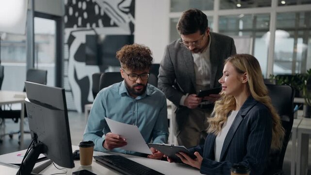 Three multiethnic business office colleagues team working together discussing work online papers data talking businessmen males with businesswoman female talk using computer digital tablet documents