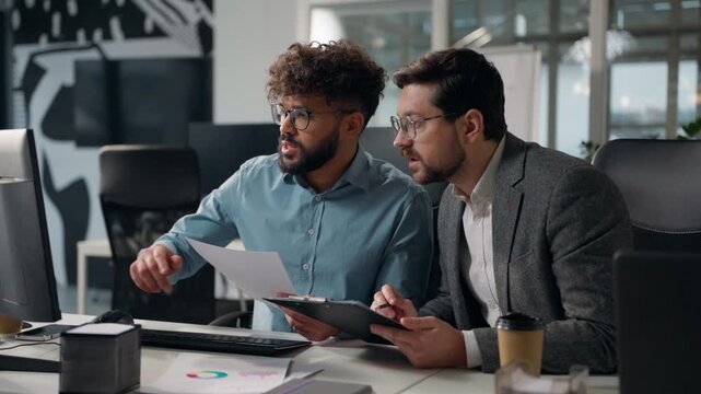Two multiracial business men working with documents computer talking holding paper checklist analyzing chart graph businessmen talk efficiency of job males review agenda diagram progress in office
