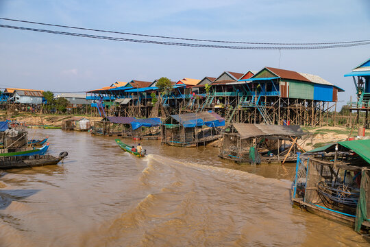 View of stilted houses reflecting in the muddy water, as a longtail boat zips along the river in Floating Village, Siem Reap Province, Cambodia.