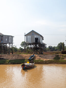 View of stilted houses mirroring in the muddy water with a small boat docked at the shore, evoking a sense of tranquility, Floating Village, Siem Reap Province, Cambodia.