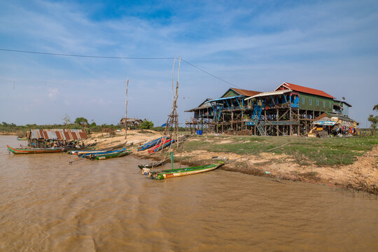 View of stilted homes rise above the muddy waters, colorful boats rest on the shore under a vast blue sky, a vibrant scene, Floating Village, Siem Reap Province, Cambodia.