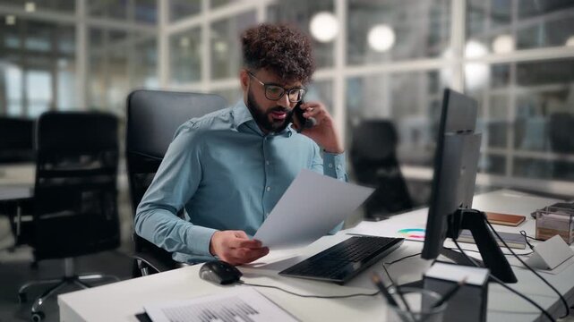 Ethnic Arabian business man working computer looking in papers report talking phone multitasking guy businessman with diagram result financial stats risk look at desktop hold document male at office