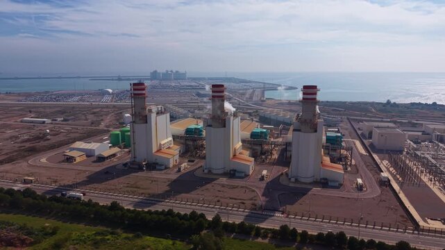 Aerial view of coastal combined cycle gas plant with gas turbines, HRSG units, steam cycle blocks, exhaust stacks and grid interconnection near port logistics hub