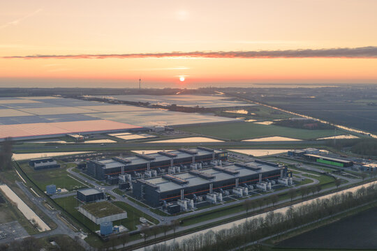 Aerial view of a modern industrial complex nestled amidst a landscape of canals and fields under a vibrant sunrise, Middenmeer, North Holland, Netherlands.
