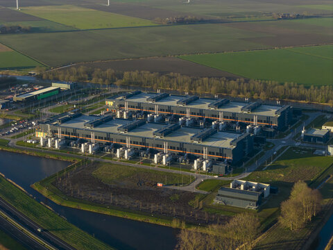 Aerial view of a stark, modern data center complex contrasts sharply with the surrounding patchwork of green fields and winding canals, Middenmeer, North Holland, Netherlands.