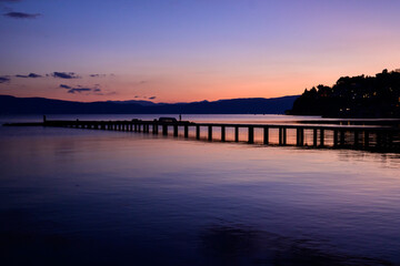 Obraz premium A long wooden pier stretches into the glassy Lake Ohrid, reflecting the stunning purple and orange twilight sky. Silhouettes of people walk along the structure.