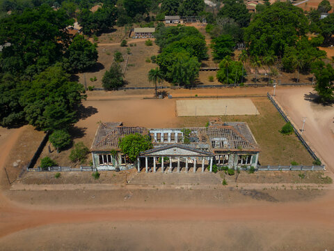 Aerial view of the ruins of a colonial building with a white colonnade and overgrown vegetation surrounded by dirt roads and lush green trees in Bijagos, Guinea Bissau.