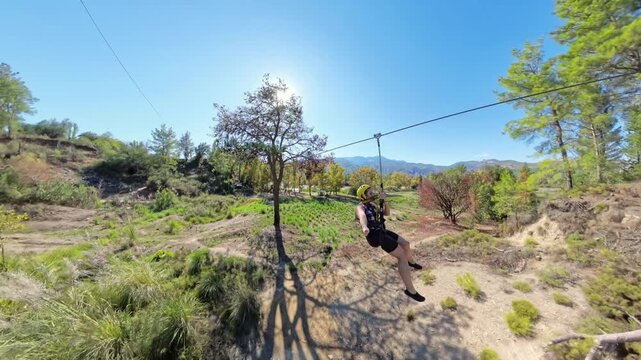 A dynamic shot of a woman ziplining through a forested area, with clear skies and mountain views in the background, wearing a helmet and harness for safety.