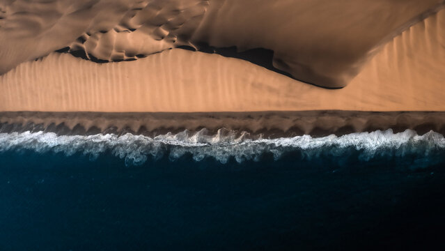 Aerial view of the dramatic meeting point between golden desert dunes and the crashing waves of the Atlantic Ocean in Walvis Bay, Erongo Region, Namibia.