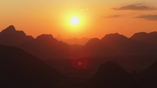 Paramotor flies over silhouetted mountains in Vang Vieng, Laos during a warm sunset with a hot air balloon appearing