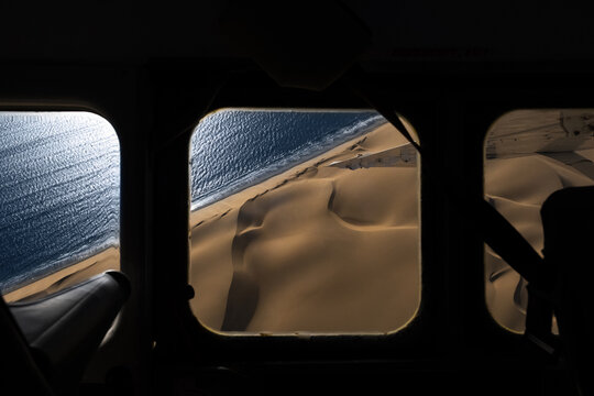Aerial view of the Namib Desert sand dunes meeting the Atlantic Ocean coastline through an airplane window in Walvis Bay, Erongo Region, Namibia.