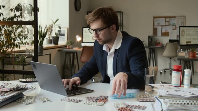Overwhelmed gen Z man fumbling for pillbox and taking pill with water while typing on laptop at white desk featuring blister packs and medication bottles in office setting