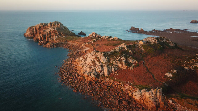 Aerial view of the rugged coastline where ochre rocks meet the turquoise sea, a landscape bathed in the warm glow of the setting sun, Plougasnou, Bretagne, France.