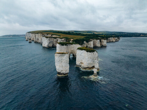 Aerial view of chalk sea stacks stand defiant against the relentless waves, crowned with verdant grass under a brooding sky, Old Harry Rocks, England, United Kingdom.