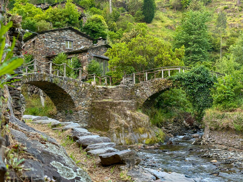 Aerial view of a stone bridge arching gracefully over a stream, connecting ancient stone houses nestled amidst verdant hills, Foz d'Egua, Arganil, Piodao, Portugal.