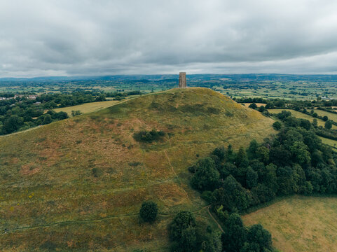 Aerial view of Glastonbury Tor crowned by a solitary tower rises majestically from the verdant landscape, a beacon under the moody sky, Glastonbury, England, United Kingdom.