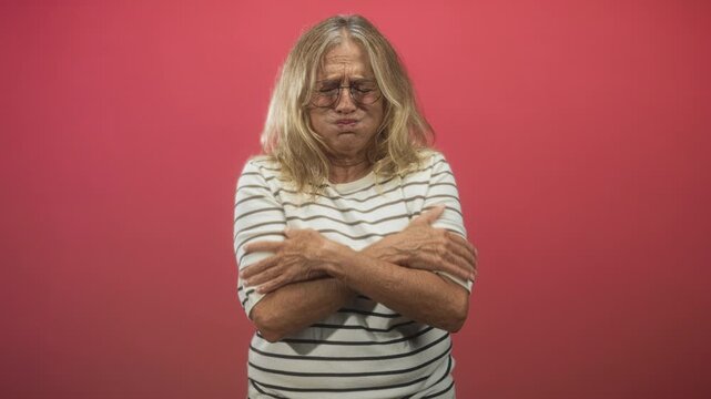 Senior woman with glasses crossing arms over chest while clutching sternum in studio with pink backdrop; pain concern.