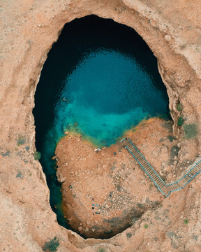 Aerial view of the Sink Hole with turquoise waters surrounded by rugged desert cliffs and a stone staircase in Dhofar Governorate, Oman.