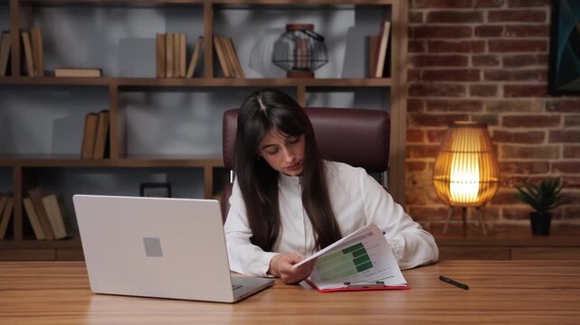 Business woman working on documents while looking at laptop in modern office setup.