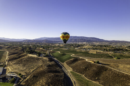 Aerial view of a vibrant hot air balloon drifts serenely over the patchwork vineyards and rolling hills under a clear blue sky, Temecula, California, United States.