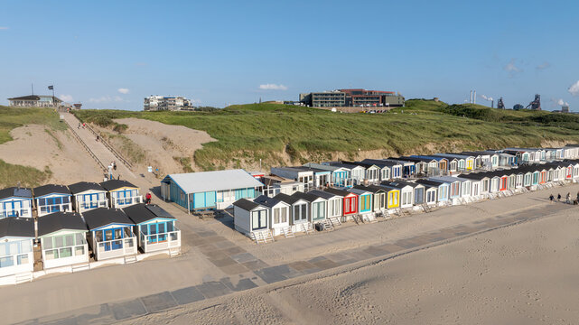 Aerial view of colourful beach huts line the sandy shore, with grassy dunes and distant industrial structures under a clear sky, Beach Wijk aan Zee, North Holland, Netherlands.