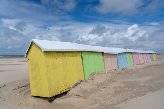 View of brightly colored beach huts stand in a row against the pale sand under a vast sky of blue and white clouds, Berck-Plage, Hauts-de-France, France.